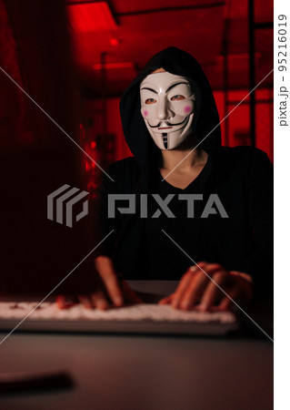 Close-up vertical shot of wanted hacker man wearing anonymous mask and sweatshirt with hood engaged in hacking into security systems, sitting in dark room with red neon lights. 95216019