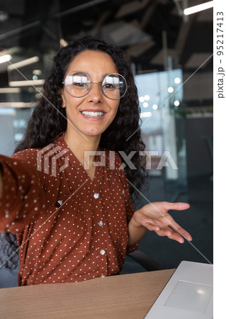 Vertical shot, video call from office hispanic business woman working inside modern office building, talking remotely with friends using app on phone for video call 95217413