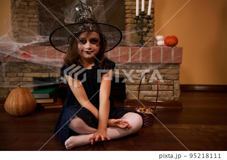 Cute little girl with Halloween themed make-up, dressed as a witch in a wizard's hat, looks at camera, sitting against the backdrop of a fireplace covered with cobwebs, pumpkins and old mystical books 95218111
