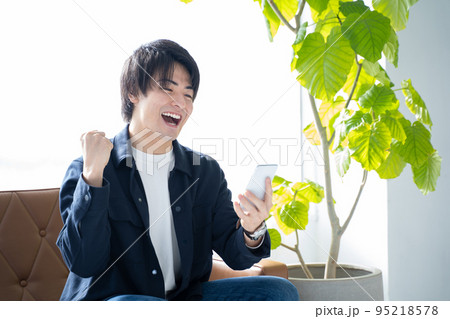 スマホを見てガッツポーズをする20代男性-リビングのソファーに座りリラックス スマホを見てガッツポーズをする20代男性-リビングのソファーに座りリラックス 95218578