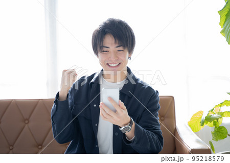 スマホを見てガッツポーズをする20代男性-リビングのソファーに座りリラックス スマホを見てガッツポーズをする20代男性-リビングのソファーに座りリラックス 95218579