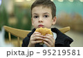 Tired sad boy of seven years old eats a burger at a food court after a school day. Child in sweatshirt enjoying a juicy burger 95219581