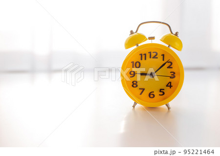A yellow alarm clock stands on a white table by the window with white curtains A yellow alarm clock stands on a white table by the window with white curtains 95221464