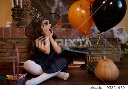 Surprised Caucasian little girl in witch black dress and wizard hat, looks at balloons, sitting near pumpkin and basket with candies against a fireplace covered with spider web. Happy Halloween party 95221970
