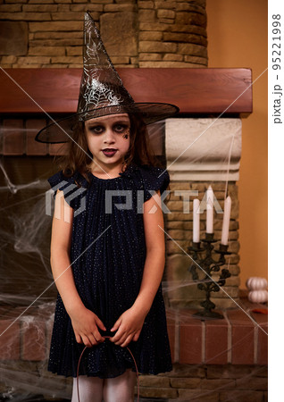 Charming little girl, a child witch in wizard hat, with a facial art makeup posing against a fireplace with spider web and candles on a candlestick. 31 October. Child celebrating Halloween. Autumn 95221998
