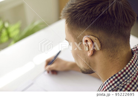Hearing impaired student wearing modern hearing aid is sitting at desk and writing 95222695