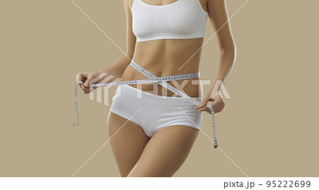 Slender woman in lingerie measures figure with tape measure standing on beige background. Slender woman in lingerie measures figure with tape measure standing on beige background. 95222699