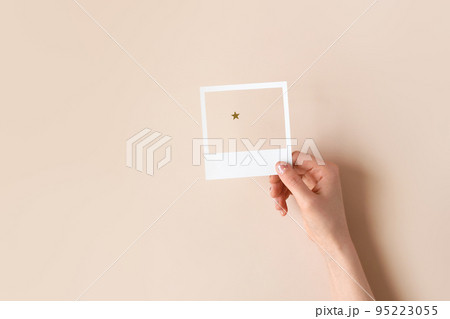 Female hand holding a white paper frame with a star seen through it on beige background. Festive minimalistic Xmas concept. Christmas postcard template. Little things matter. Birthday theme design 95223055