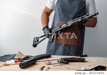 Close up of young man in apron disassembling a gun above the table Close up of young man in apron disassembling a gun above the table 95223850