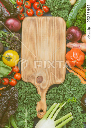 Organic vegetables assortment: broccoli cauliflower carrots tomatoes kale pak choy onions. Healthy local farm produce on table surrounding wood chopping board, top view, selective focus Organic vegetables assortment: broccoli cauliflower carrots tomatoes kale pak choy onions. Healthy local farm produce on table surrounding wood chopping board, top view, selective focus 95223945