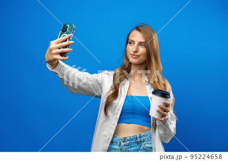 Portrait of smiling beautiful woman with takeout coffee using smartphone against blue background in studio Portrait of smiling beautiful woman with takeout coffee using smartphone against blue background in studio 95224658
