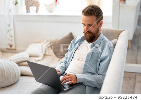 Young bearded man sitting on sofa at home using laptop computer Young bearded man sitting on sofa at home using laptop computer 95225474