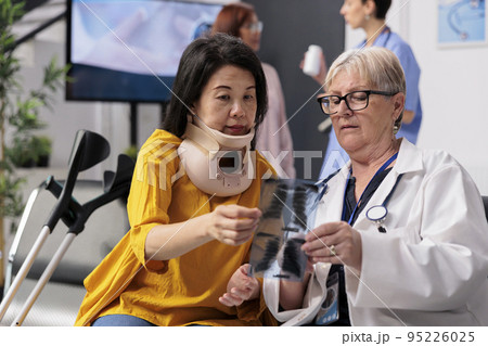 Physician consulting patient with cervical neck collar in hospital waiting room, looking at lungs xray exam, Senior medic and patient discussing about healthcare at checkup visit 95226025