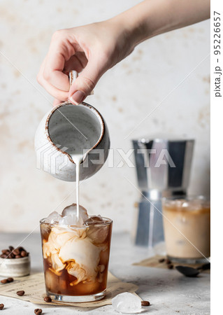 Pouring milk in coffee with ice cubes with beautiful twists on light background. Iced latte coffee still life 95226657