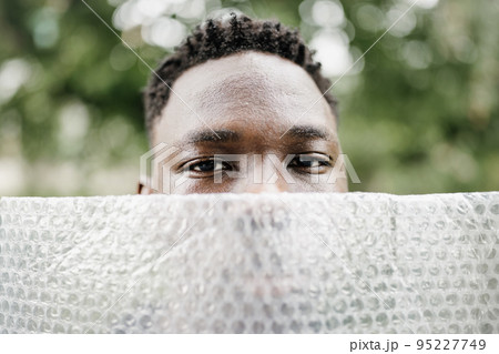 Young African American man outdoor, close-up street portrait with plastic bubble wrap foil. Ecology, environmental conversation, mental health concept. Fall or spring season Young African American man outdoor, close-up street portrait with plastic bubble wrap foil. Ecology, environmental conversation, mental health concept. Fall or spring season 95227749