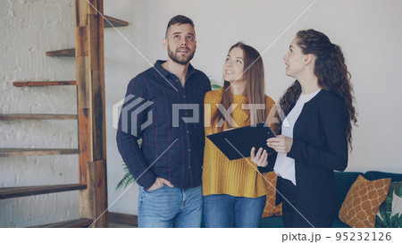 Cheerful house agent is talking to buyers adorable young couple discussing conditions of sale and showing papers. Smiling clients are excited about new apartment. 95232126
