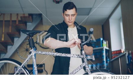 Strong handsome man skilled mechanic is concentrated on checking and repairing handlebar of broken bicycle in his small workshop. Skilled people and maintenance concept. 95232127