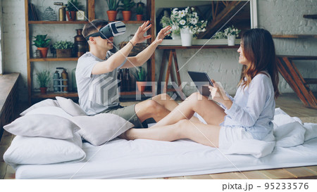 Young man is using virtual reality glasses while his attractive wife is touching tablet screen, looking at her husband and smiling. Modern technology and human relationship concept. 95233576