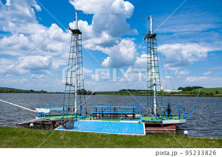 A homemade wooden boat with masts on iron barrels for fishing is moored to the shore of the lake. A homemade wooden boat with masts on iron barrels for fishing is moored to the shore of the lake. 95233826