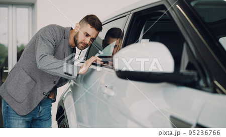 Stylish bearded guy is choosing new automobile walking around luxury car and touching it in car dealership. Rich people, transportation and business class vehicles concept. Stylish bearded guy is choosing new automobile walking around luxury car and touching it in car dealership. Rich people, transportation and business class vehicles concept. 95236766