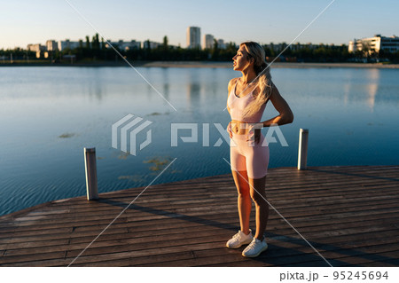 Sporty young woman exercising and stretching neck for warming up before running or working out near water on background of cityscape. Blonde sportswoman exercising in soft sunlight of summer morning. Sporty young woman exercising and stretching neck for warming up before running or working out near water on background of cityscape. Blonde sportswoman exercising in soft sunlight of summer morning. 95245694