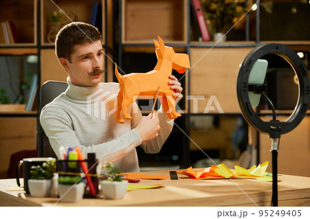 Young man sitting at home, holds a master class on origami and recording vlog at home studio. Hobbies and leisure activities. Remote training, online classes 95249405