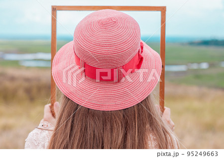 Girl in pink straw hat holds empty wooden picture frame in hands on nature landscape background faceless. Creative idea. 95249663