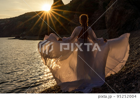 A mysterious female silhouette with long braids stands on the sea beach with mountain views, Sunset rays shine on a woman. Throws up a long white dress, a divine sunset. A mysterious female silhouette with long braids stands on the sea beach with mountain views, Sunset rays shine on a woman. Throws up a long white dress, a divine sunset. 95252004