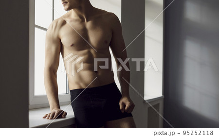 Young man with perfect athletic body in underpants and with naked torso poses by window. Young man with perfect athletic body in underpants and with naked torso poses by window. 95252118