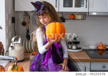 Child decorates the kitchen in home for Halloween. Girl in a witch costume plays with the decor for the holiday - bats, jack lantern, pumpkins. Autumn comfort in house, Scandi-style kitchen, loft 95252576