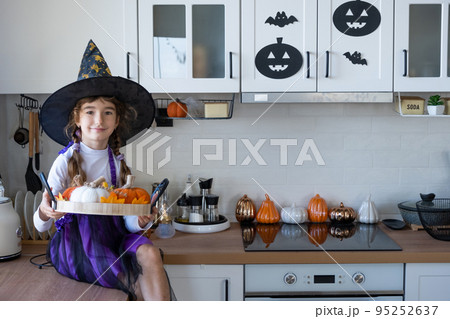 Child decorates the kitchen in home for Halloween. Girl in a witch costume plays with the decor for the holiday - bats, jack lantern, pumpkins. Autumn comfort in house, Scandi-style kitchen, loft Child decorates the kitchen in home for Halloween. Girl in a witch costume plays with the decor for the holiday - bats, jack lantern, pumpkins. Autumn comfort in house, Scandi-style kitchen, loft 95252637