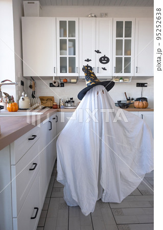 A child in sheets with cutout for eyes like a ghost costume in the kitchen decorated for the Halloween holiday. A kind little funny ghost. Halloween Party A child in sheets with cutout for eyes like a ghost costume in the kitchen decorated for the Halloween holiday. A kind little funny ghost. Halloween Party 95252638