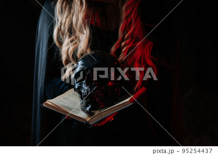 Witch in black costume with black magic book. Woman holds skull in hands. Death, spiritual rituals 95254437