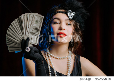 Rich luxurious woman dressed in Art Deco style waves a bundle of dollars banknotes like fan. Vintage 95255016