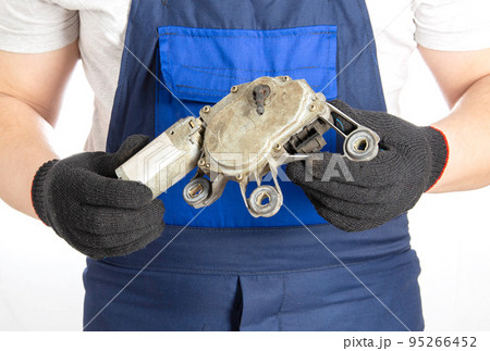 A car mechanic holds a broken electric windshield wiper motor in his hands against the background of a blue overalls. Repair and replacement of car parts in a car service, close-up 95266452