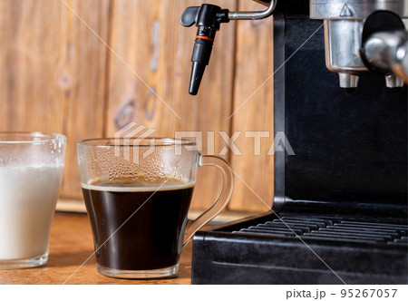 A cup of black coffee and a cup of milk stand near a coffee maker with a manual cappuccino maker. Wood background, close-up 95267057