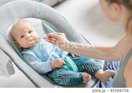Mother spoon feeding her baby boy infant child in baby chair with fruit puree. Baby solid food introduction concept. 95268738