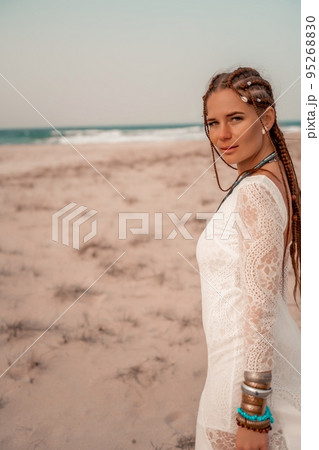 Model in boho style in a white long dress and silver jewelry on the beach. Her hair is braided, and there are many bracelets on her arms. 95268830