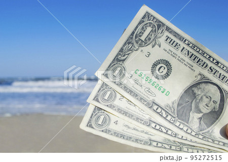 Person holding three paper dollar bills on background of sea and sea waves with white foam, sandy beach and clear blue sky on sunny summer day. Concept of money, expenses on vacation, travel, tourism 95272515