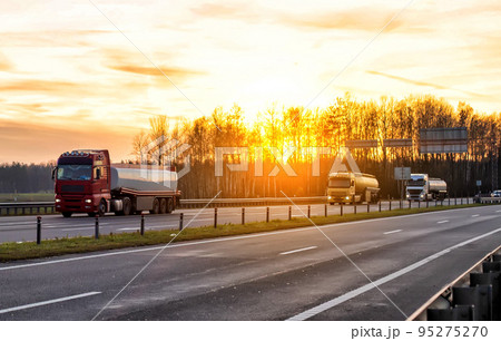 A convoy of three trucks transports dangerous goods in tank semi-trailers on a motorway against the backdrop of sunset in the evening. A convoy of three trucks transports dangerous goods in tank semi-trailers on a motorway against the backdrop of sunset in the evening. 95275270