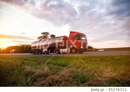 A modern truck with a semi-trailer tanker transports dangerous goods against the backdrop of a sunset in summer. Liquid cargo transportation. Copy space for text A modern truck with a semi-trailer tanker transports dangerous goods against the backdrop of a sunset in summer. Liquid cargo transportation. Copy space for text 95276111