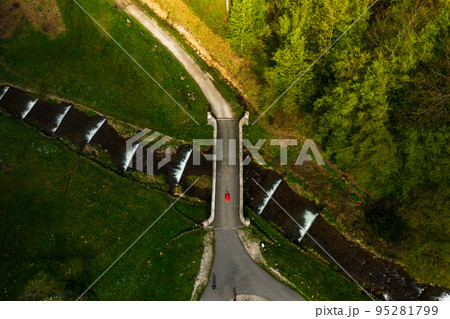 Stone arched bridge in the park, pedestrian bridge over the cascading river, woman lying on the bridge, aerial view. Stone arched bridge in the park, pedestrian bridge over the cascading river, woman lying on the bridge, aerial view. 95281799