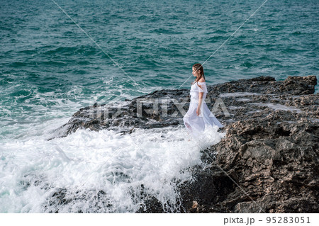 A woman stands on a rock in the sea during a storm. Dressed in a white long dress, the waves break on the rocks and white spray rises. 95283051