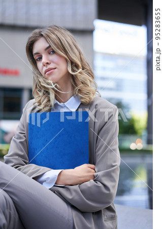 Corporate woman holding documents in hands, sitting on a street, wearing office suit, looking confident at camera, carry a folder with work Corporate woman holding documents in hands, sitting on a street, wearing office suit, looking confident at camera, carry a folder with work 95283655
