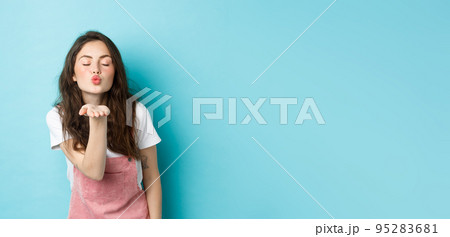 Portrait of lovely, beautiful brunette woman sending air kiss at camera, blowing mwah with palm near puckered lips and closed eyes, standing over blue background 95283681