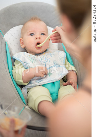 Mother spoon feeding her baby boy infant child in baby chair with fruit puree. Baby solid food introduction concept. 95284324