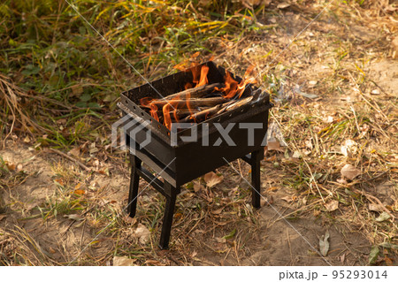 Mangal with fire outdoors. Small grill, barbecue, bbq with flame. Mini folding portable brazier filled with firewood and fire on autumn ground, hiking and picnic. Diagonal snapshot, close-up 95293014