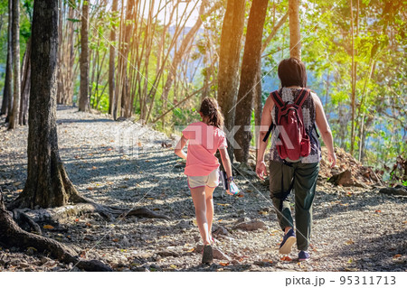 Back view of young girl and her mother walking together in forest nature path walk on trail woods background at Hellfire Pass in Kanchanaburi Thailand. Happy people relaxing on active outdoor activity 95311713