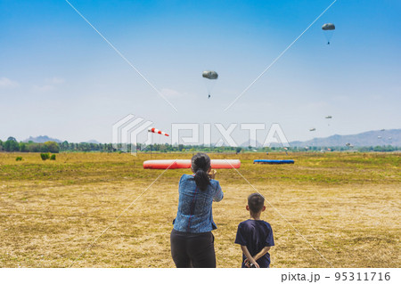Parents take video clips and photo with smart phone and watch with worry and concern during parachute training from airplane for army cadet with blurred image of parachute and landscape in background. Parents take video clips and photo with smart phone and watch with worry and concern during parachute training from airplane for army cadet with blurred image of parachute and landscape in background. 95311716