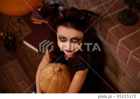 Top view Adorable gothic child, a little girl witch, enchantress, sorceress with face art makeup for Halloween party, hugging a pumpkin, smiles at camera, surrounded by a sorcery book and spider webs Top view Adorable gothic child, a little girl witch, enchantress, sorceress with face art makeup for Halloween party, hugging a pumpkin, smiles at camera, surrounded by a sorcery book and spider webs 95315172